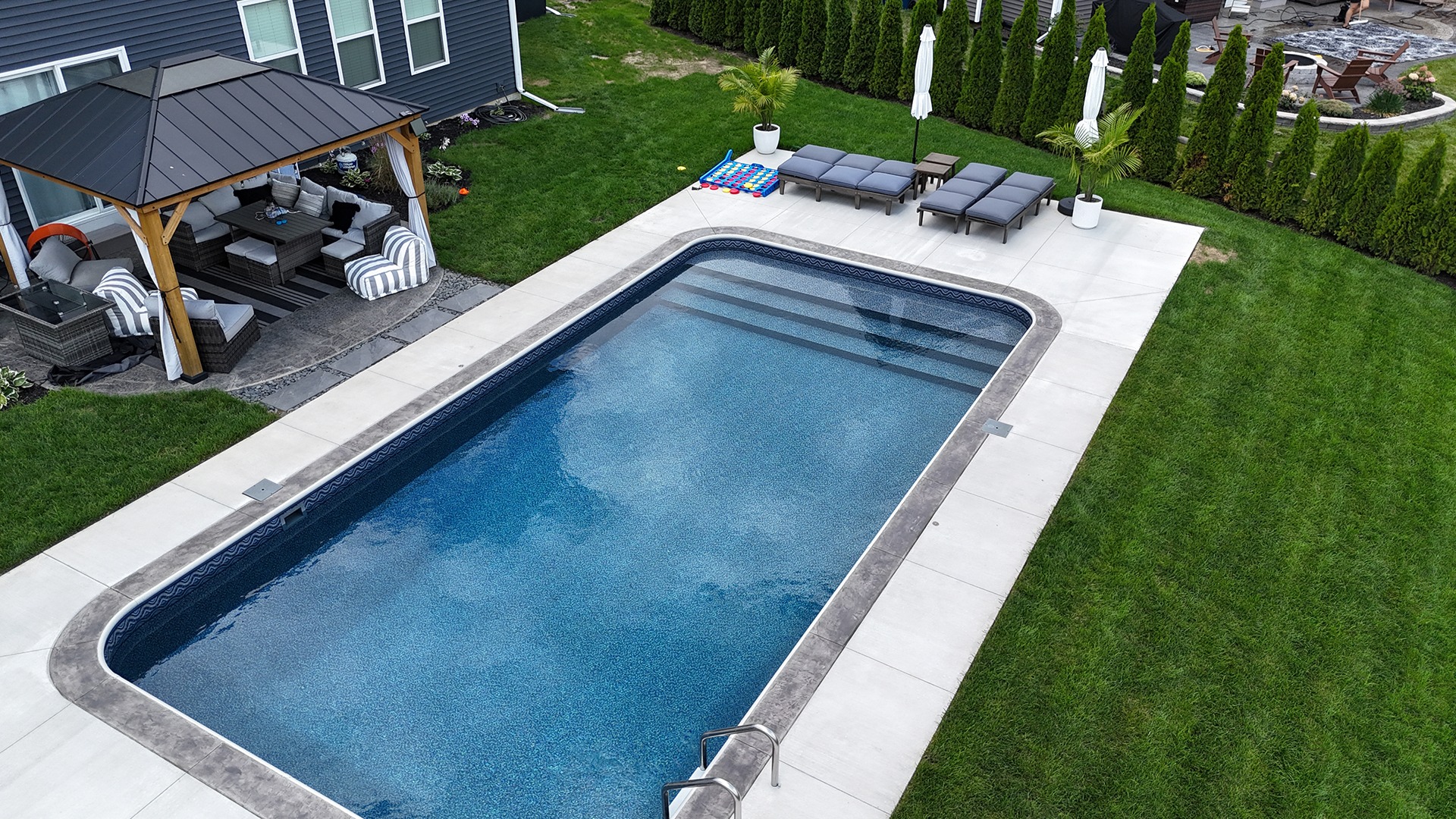 A backyard featuring a pool, patio with seating, lounge chairs, and green lawn. Umbrellas and plants enhance the comfortable outdoor setting.