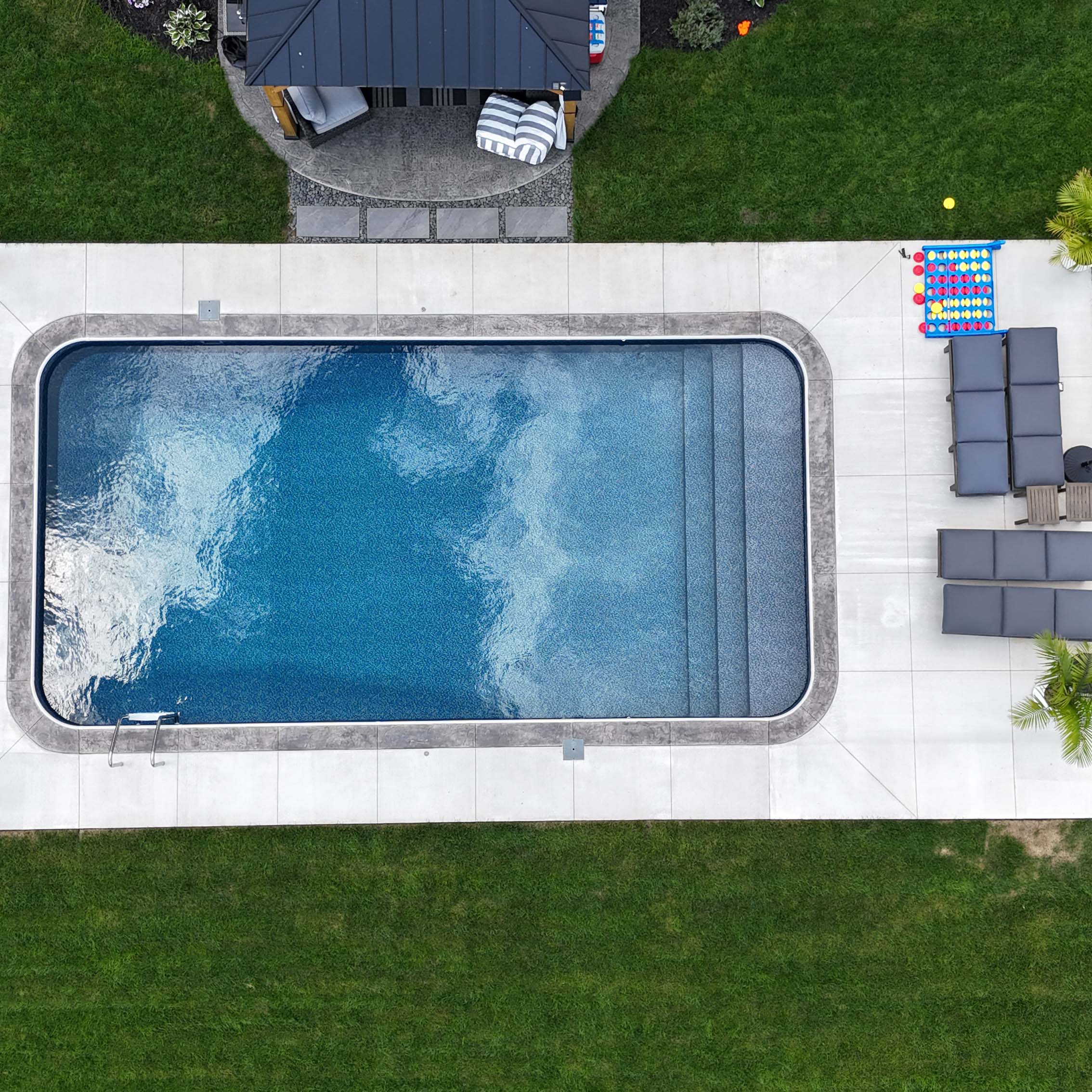 Aerial view of a backyard with a rectangular swimming pool, gray patio furniture, lush grass, and a small shaded structure.