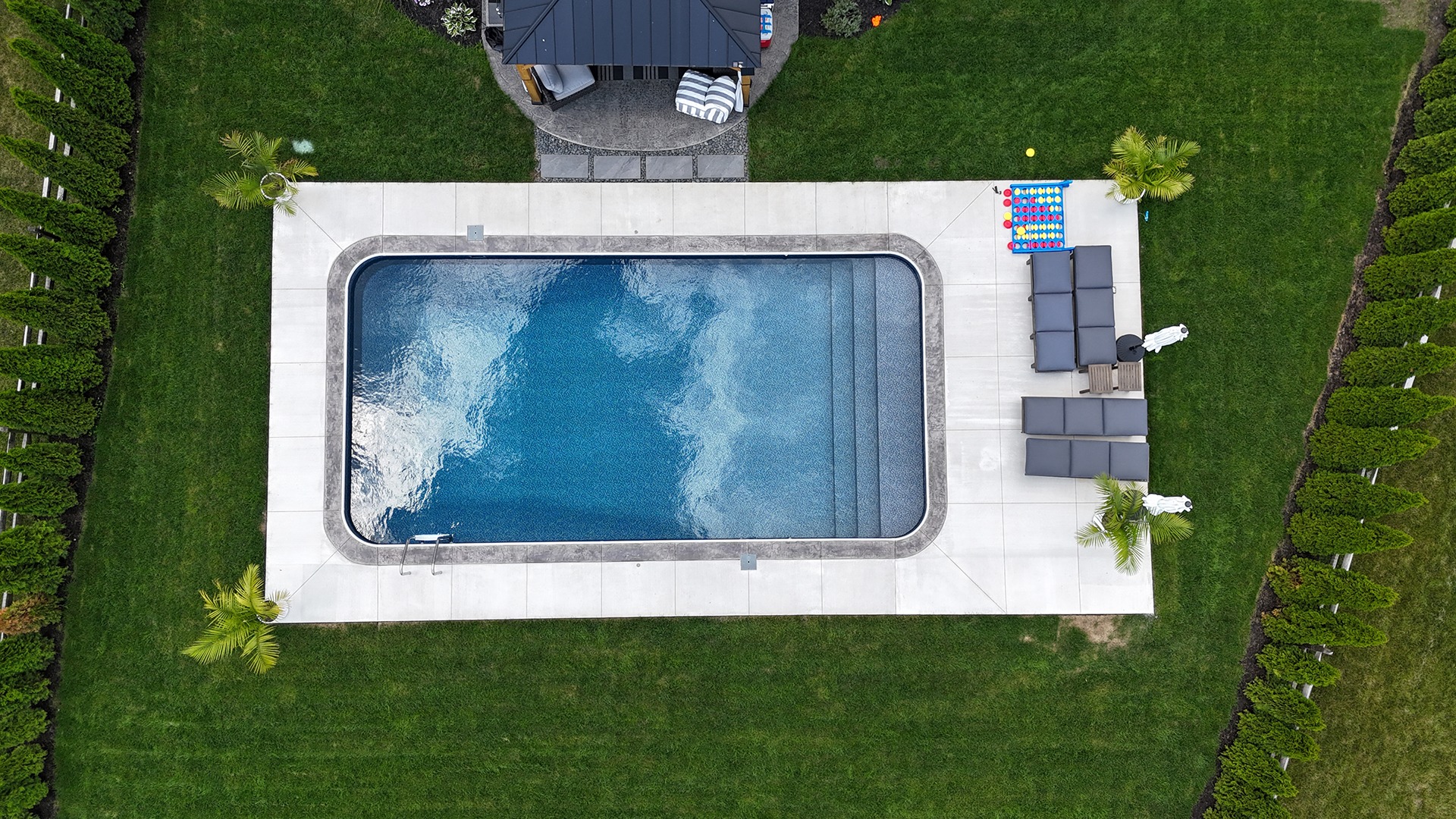 Aerial view of a rectangular swimming pool, surrounded by green lawn, patio chairs, a gazebo, and bordered by hedges and palm trees.