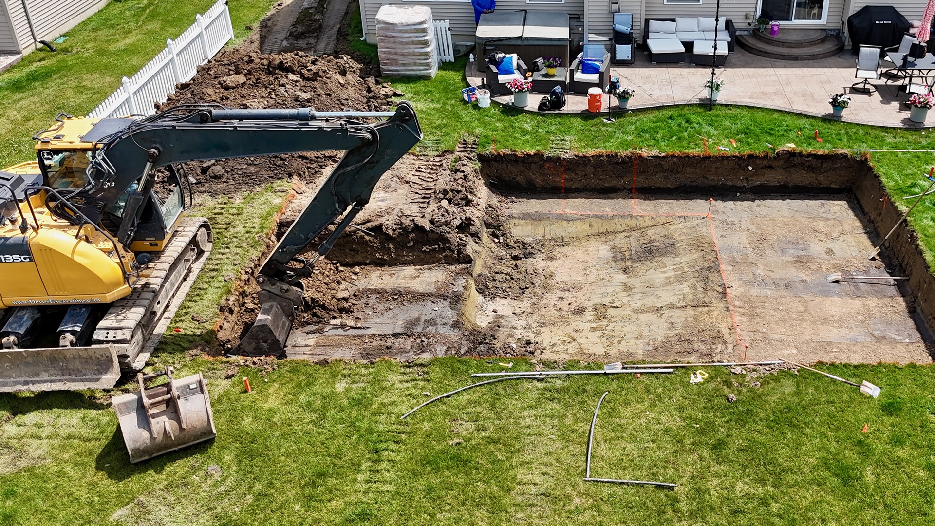 An excavator digs a large rectangular hole in a backyard, surrounded by grass, patio furniture, and a house.