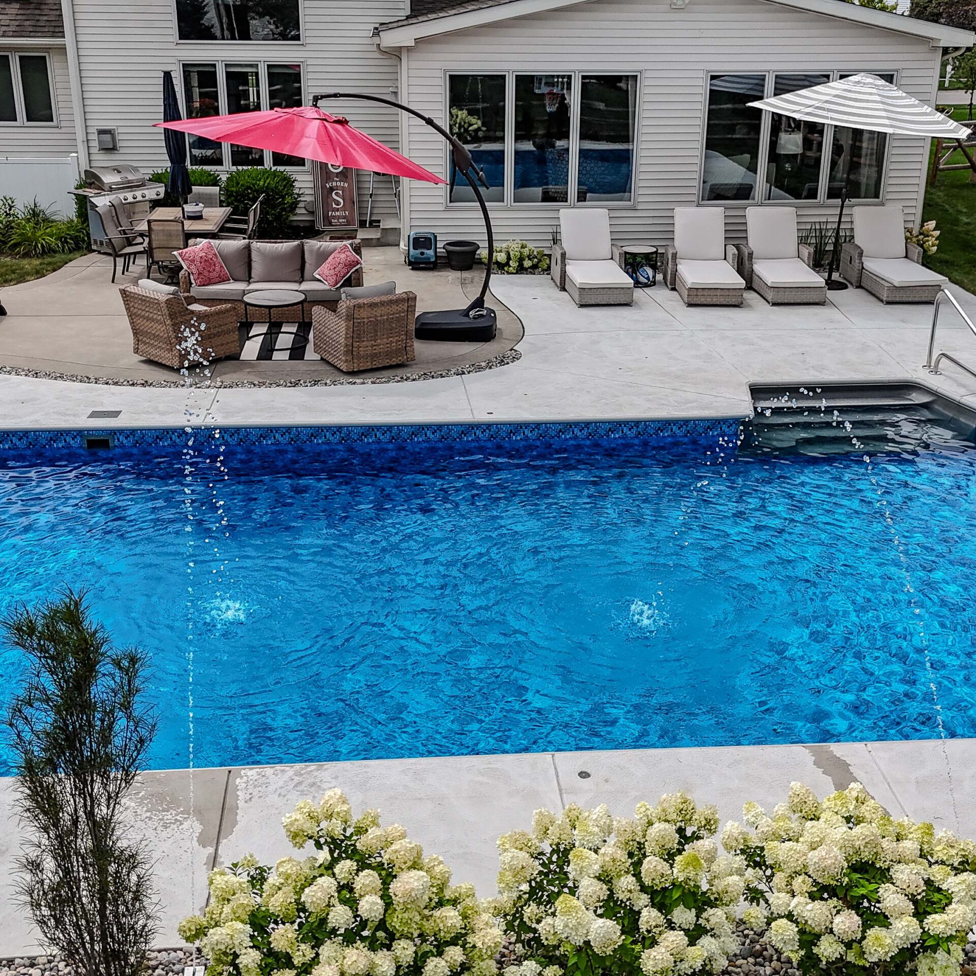 A backyard with a pool, patio furniture, red umbrella, white loungers, and blooming bushes, set against a modern house exterior.