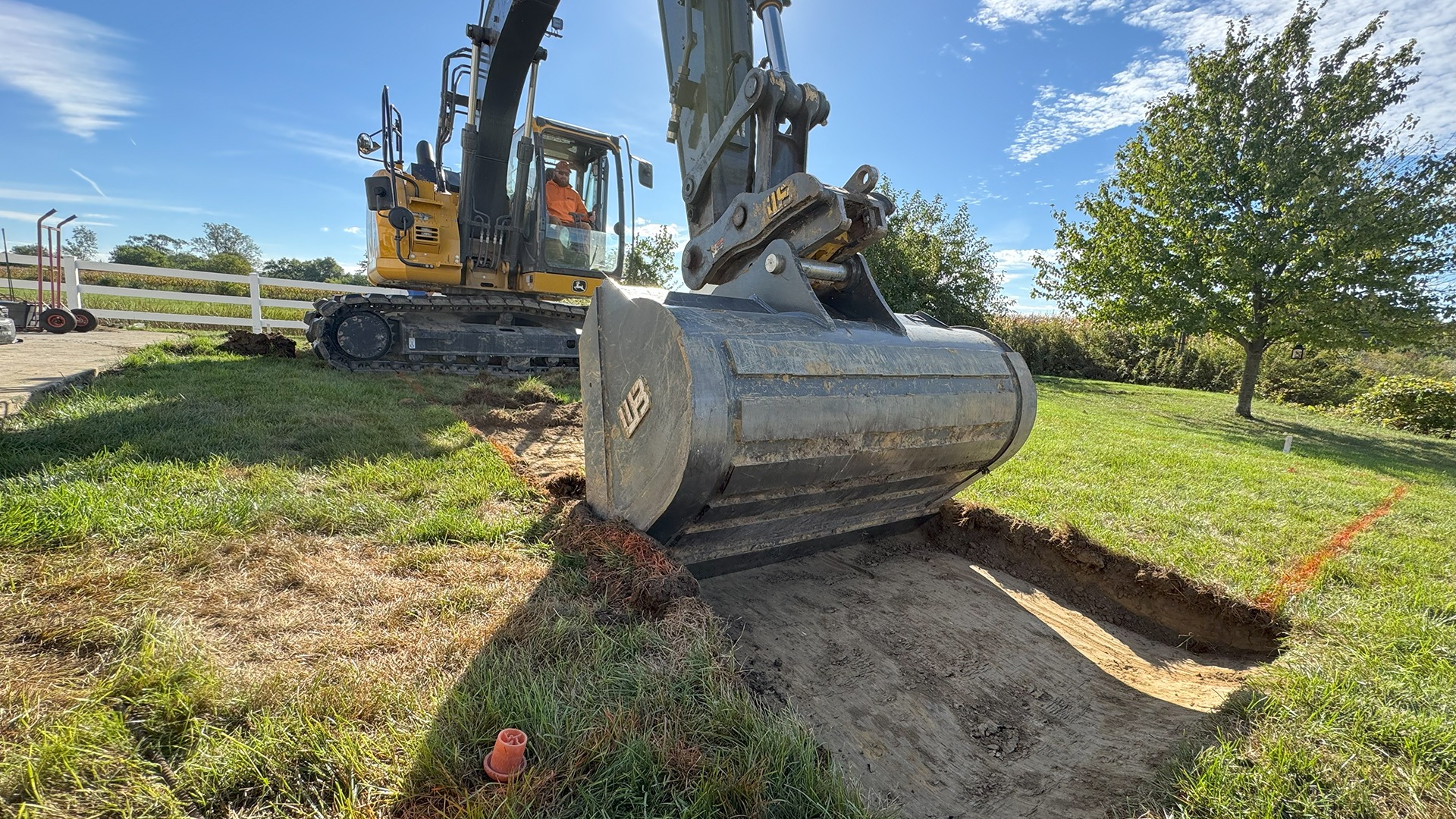 A person operates a yellow excavator, digging on a grassy field under a clear blue sky with trees and white fencing nearby.