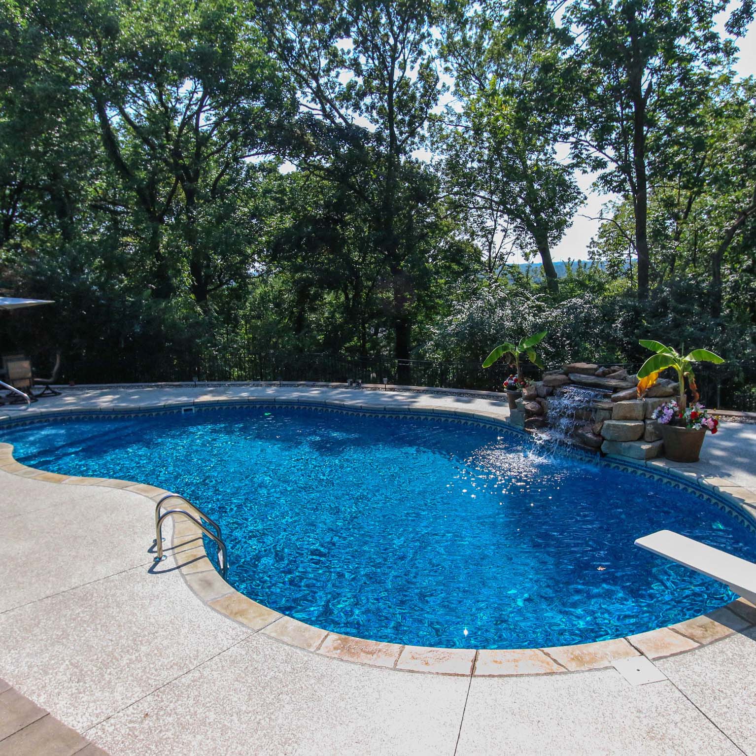 A tranquil swimming pool surrounded by lush trees, with a small waterfall feature, under a clear blue sky. No people visible.