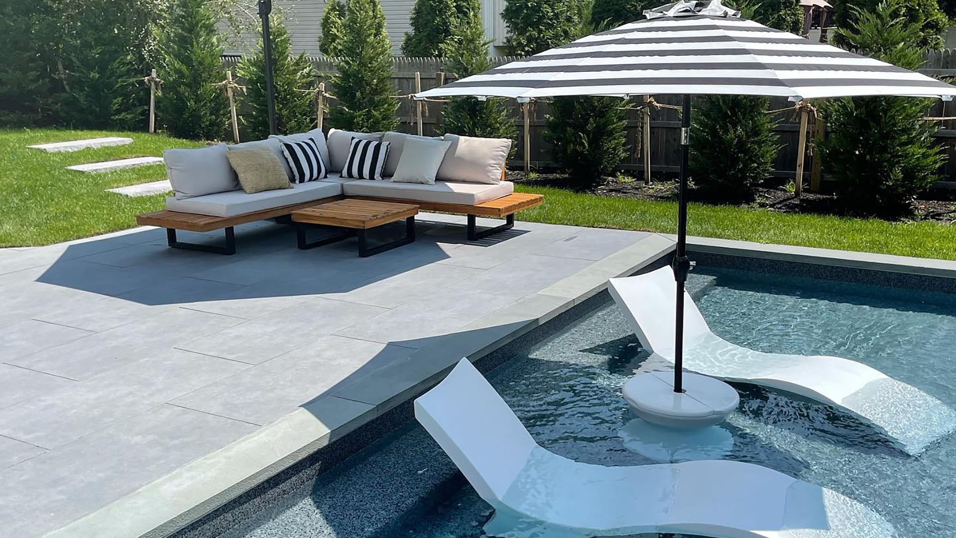 Modern backyard with pool, lounge chairs, striped umbrella, and a cozy seating area on a stone patio surrounded by lush greenery.