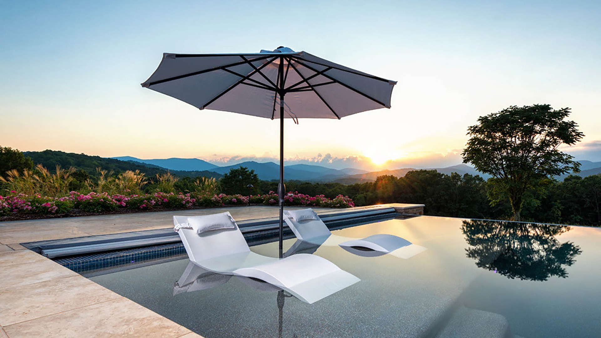 Infinity pool with submerged loungers under an umbrella, overlooking a serene mountain landscape at sunset, surrounded by lush greenery and flowers.