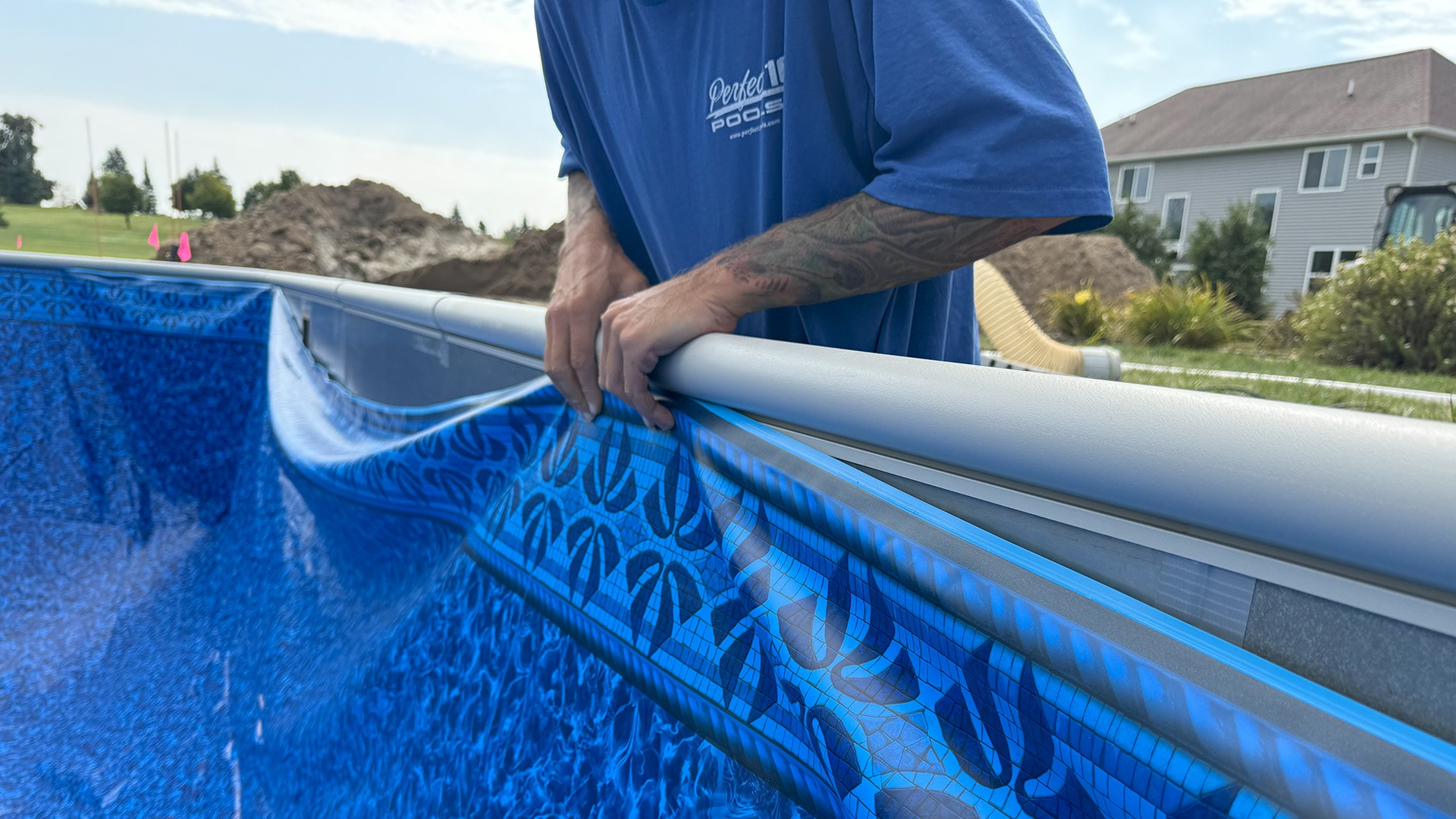 A person in a blue shirt is assembling a pool liner outdoors, with houses and a dirt pile in the background.