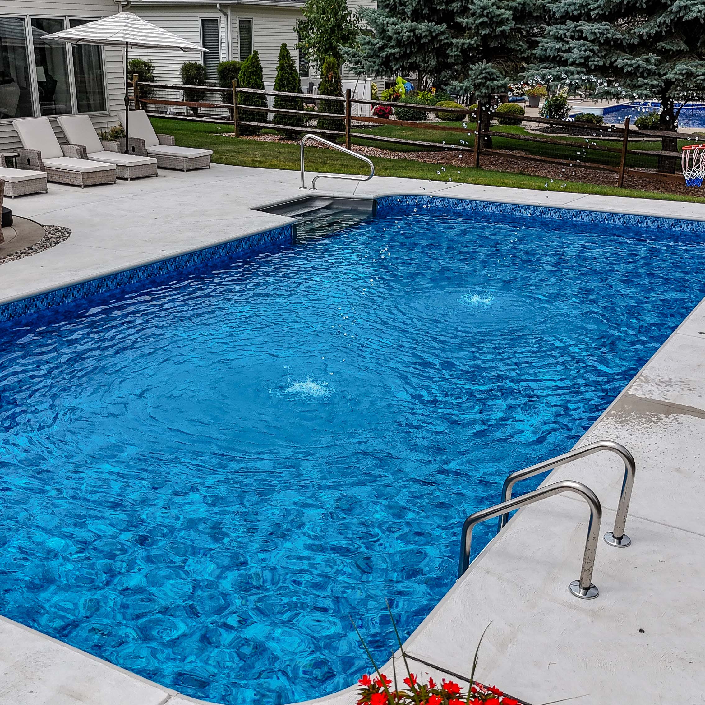 A backyard scene with a clear blue swimming pool, lounge chairs, and landscaped garden. A person in the distance, trees, and umbrella visible.