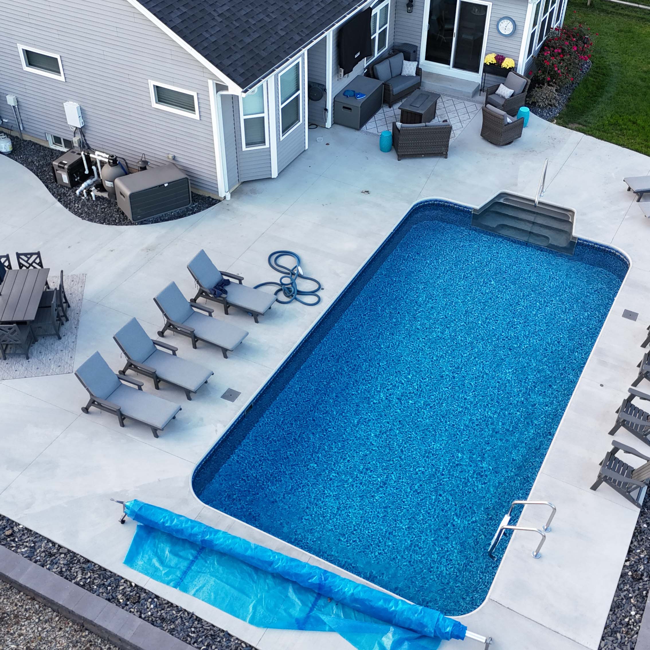 Modern backyard with rectangle pool, surrounded by lounge chairs and outdoor furniture, adjacent to a house with a gray exterior and small garden.