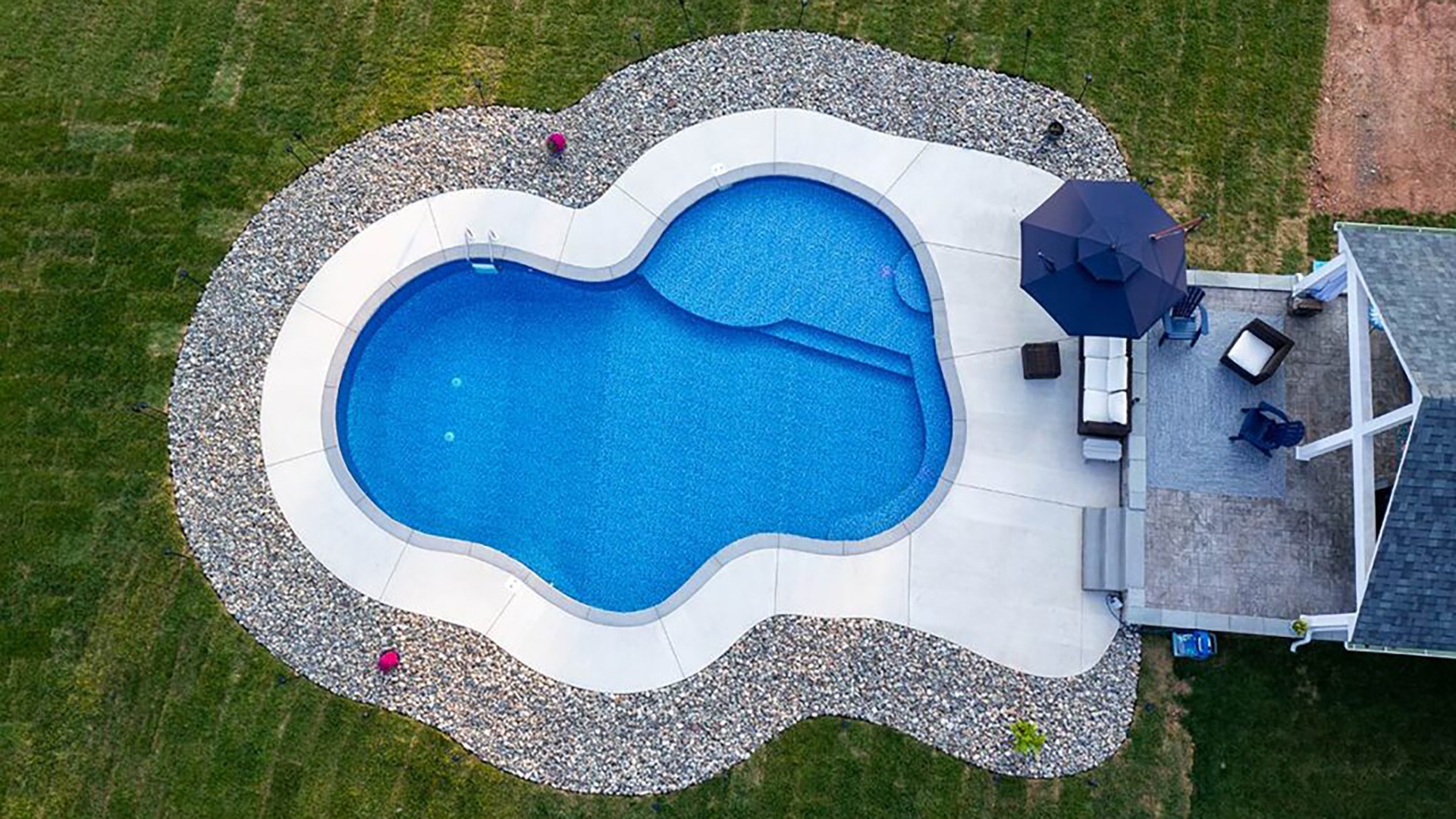 Aerial view of a uniquely shaped pool surrounded by grass, patio furniture, and an umbrella. Stone border adds a decorative touch.