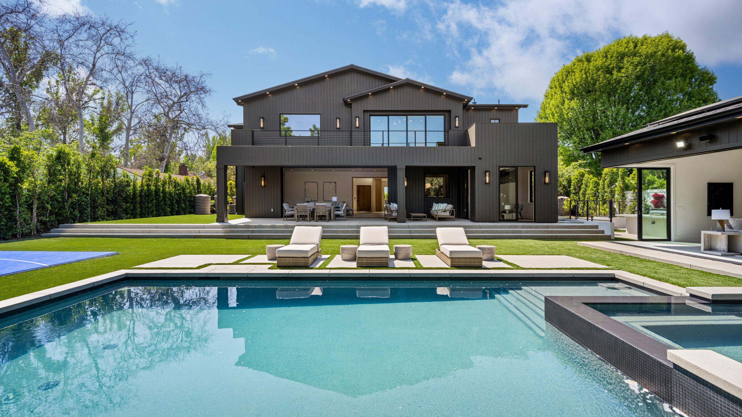 Modern house with large windows and patio, pool in foreground, surrounded by greenery and trees under a clear blue sky.