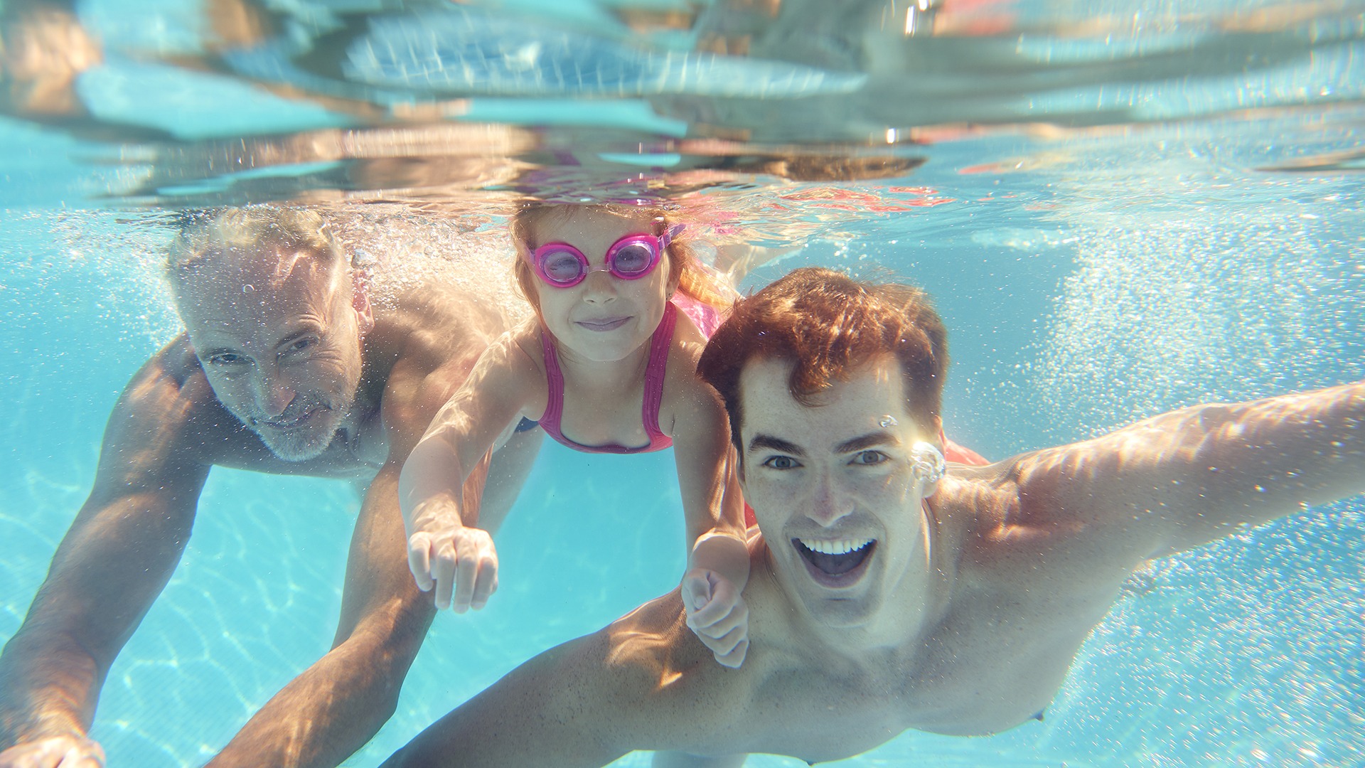 A child with pink goggles smiles underwater between two people, swimming in a clear pool, with sunlight reflecting on the surface.