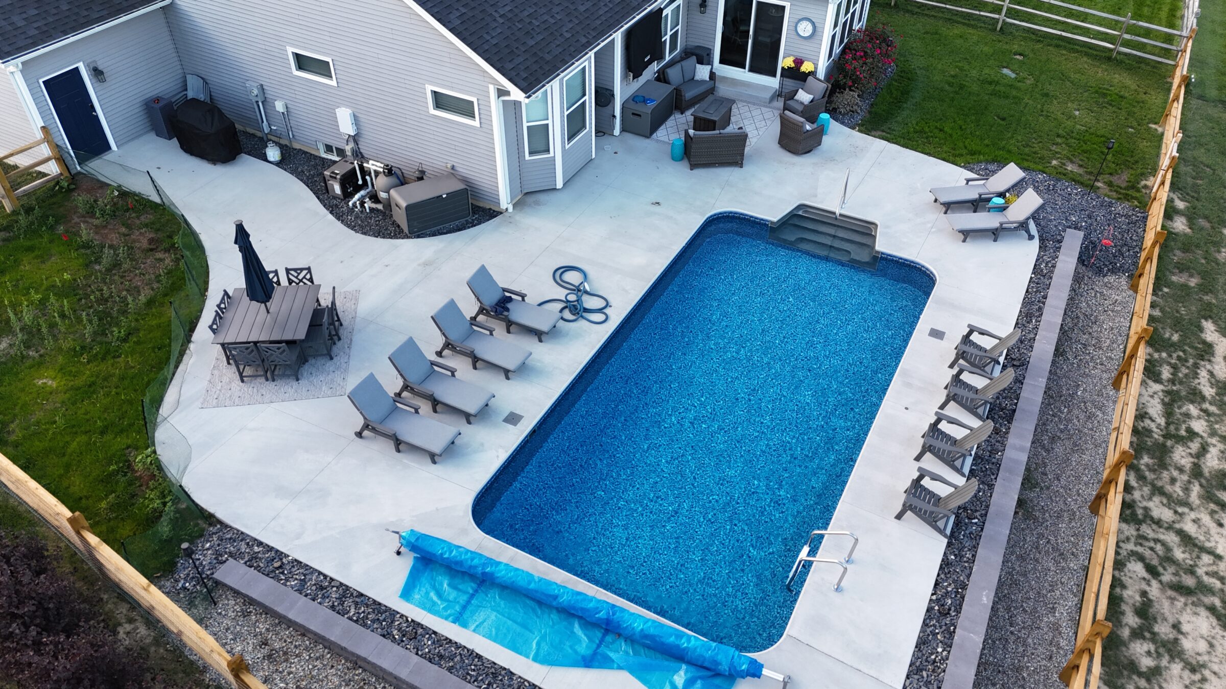 Aerial view of a rectangular vinyl liner backyard pool with light concrete decking, several loungers, outdoor seating, and a covered patio area, all enclosed by a privacy fence and landscaped with gravel accents.