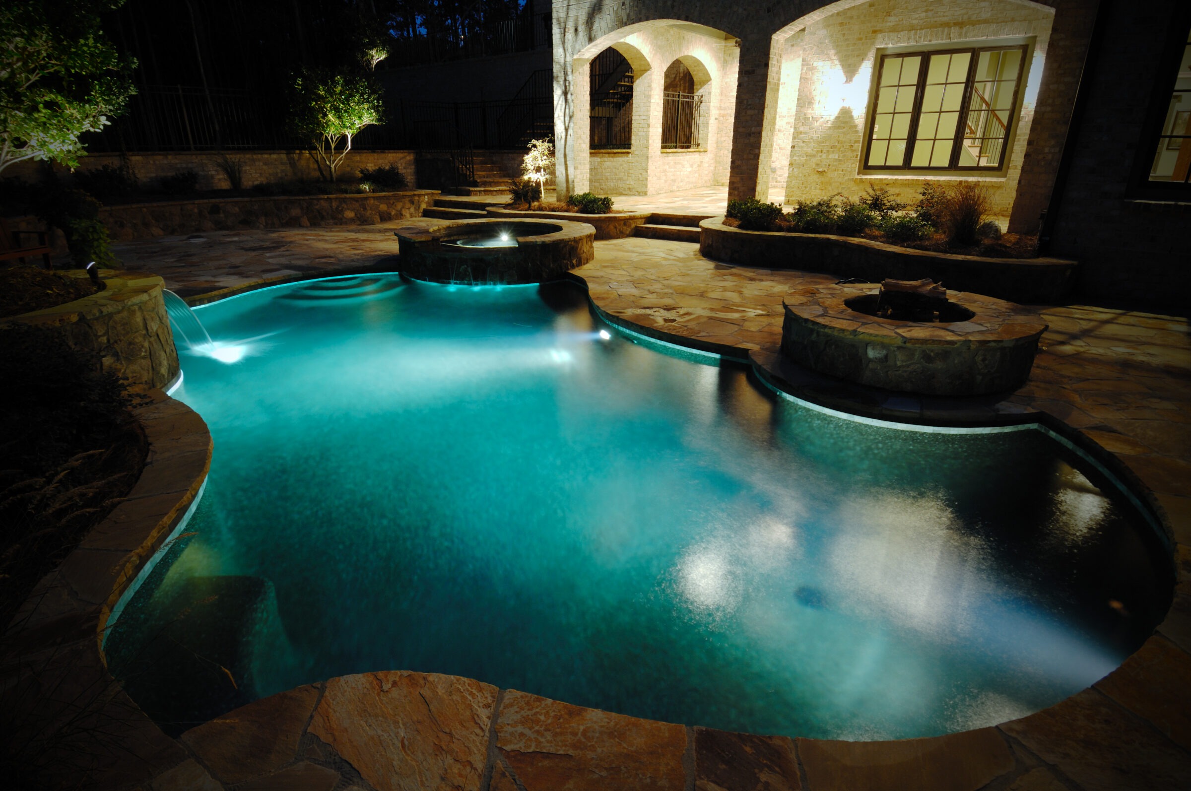 Luxury backyard vinyl liner pool illuminated at night with a glowing spa and stone fire pit, surrounded by natural stone decking and soft landscaping.