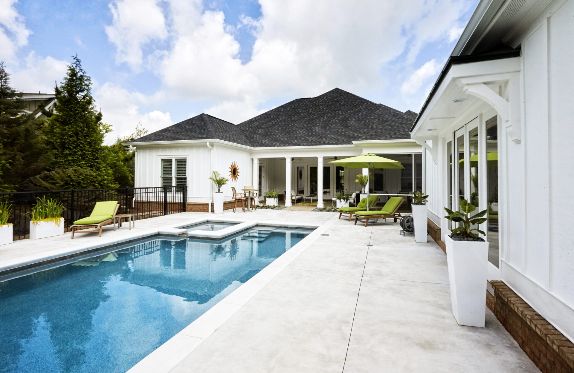 Modern backyard featuring a rectangular fiberglass pool with a built-in spa and tanning ledge, surrounded by smooth concrete decking, green cushioned loungers, and a white house with a shaded patio area.
