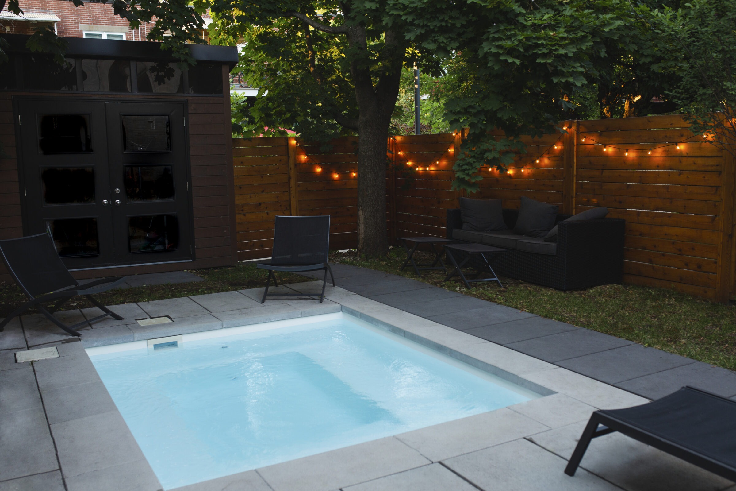 Cozy backyard plunge pool surrounded by modern black lounge chairs, a wicker loveseat, a wooden privacy fence with string lights, and mature trees providing shade and ambiance.