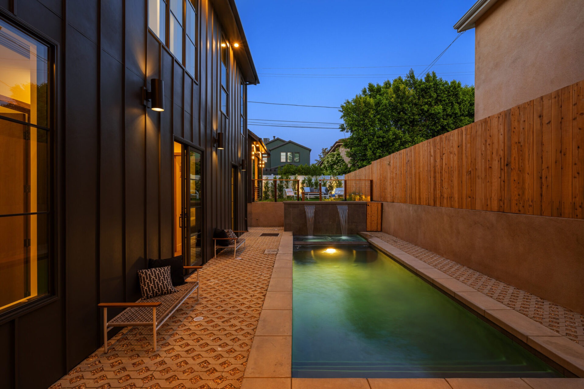 Modern narrow backyard pool illuminated at dusk, featuring two water spouts, geometric tile patio, sleek black exterior walls, and a tall wooden privacy fence.