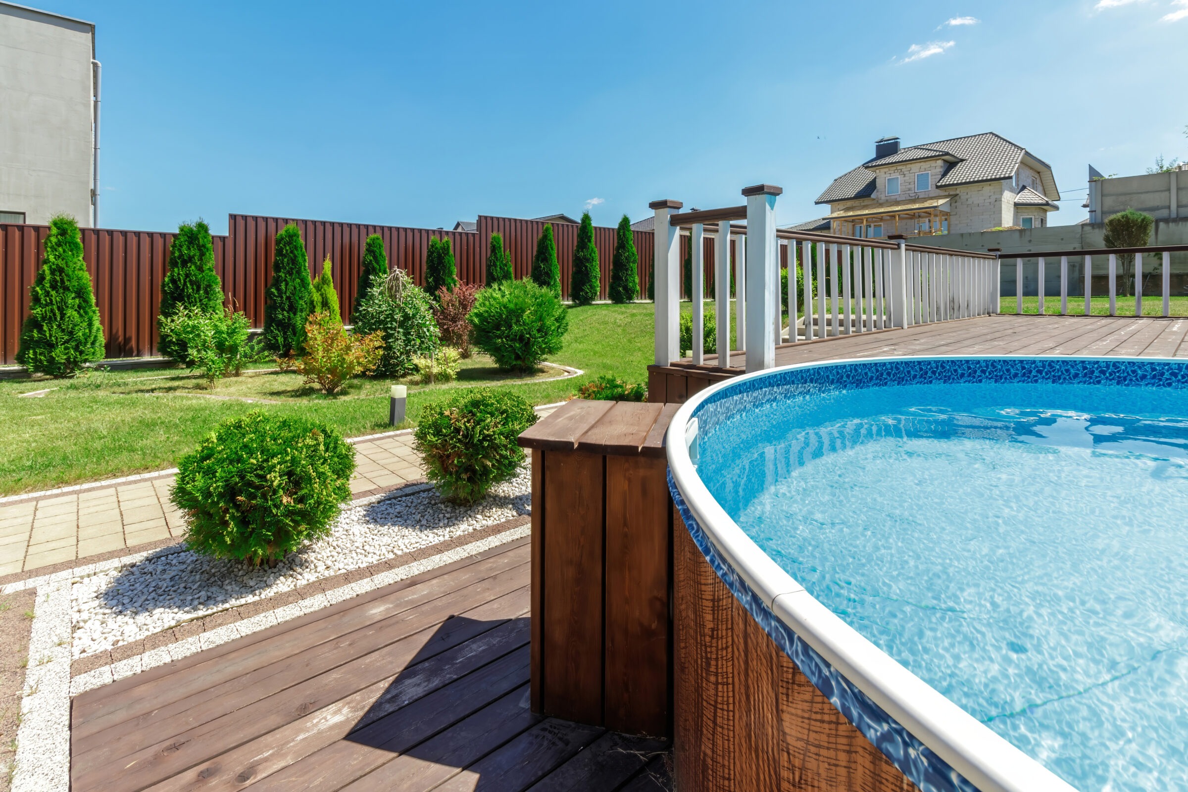 Above-ground pool with a wooden deck and white railing in a neatly landscaped backyard featuring paved walkways, decorative gravel, and small evergreen shrubs, all enclosed by a tall red privacy fence and backed by a large suburban house.