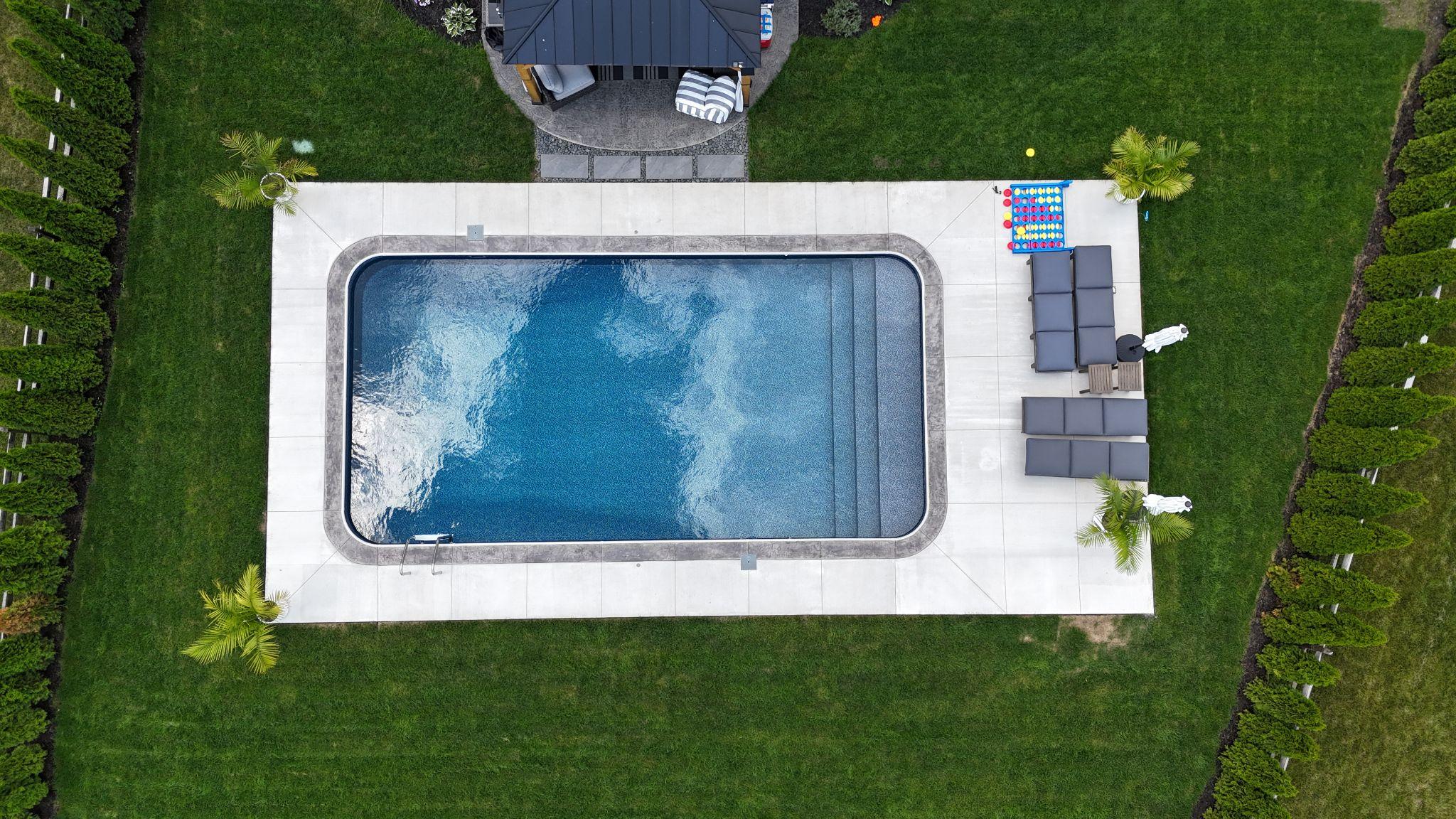 Aerial view of a rectangular backyard pool with steps leading into the shallow end. The pool is framed by a wide concrete deck, with a modern seating area and green lawn surrounding it.