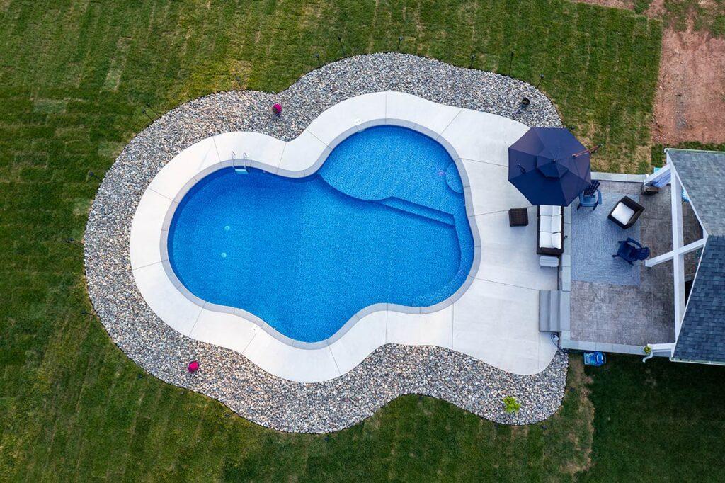 Overhead shot of a kidney-shaped pool outlined by a white concrete deck and decorative rock border. A shaded patio with outdoor seating and umbrella is positioned on one side.